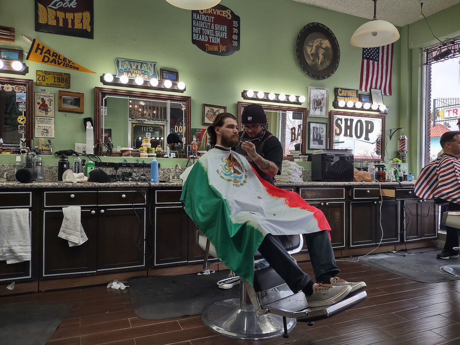 Barber trimming a client's beard, client draped in a Mexican flag cape in a vintage barbershop