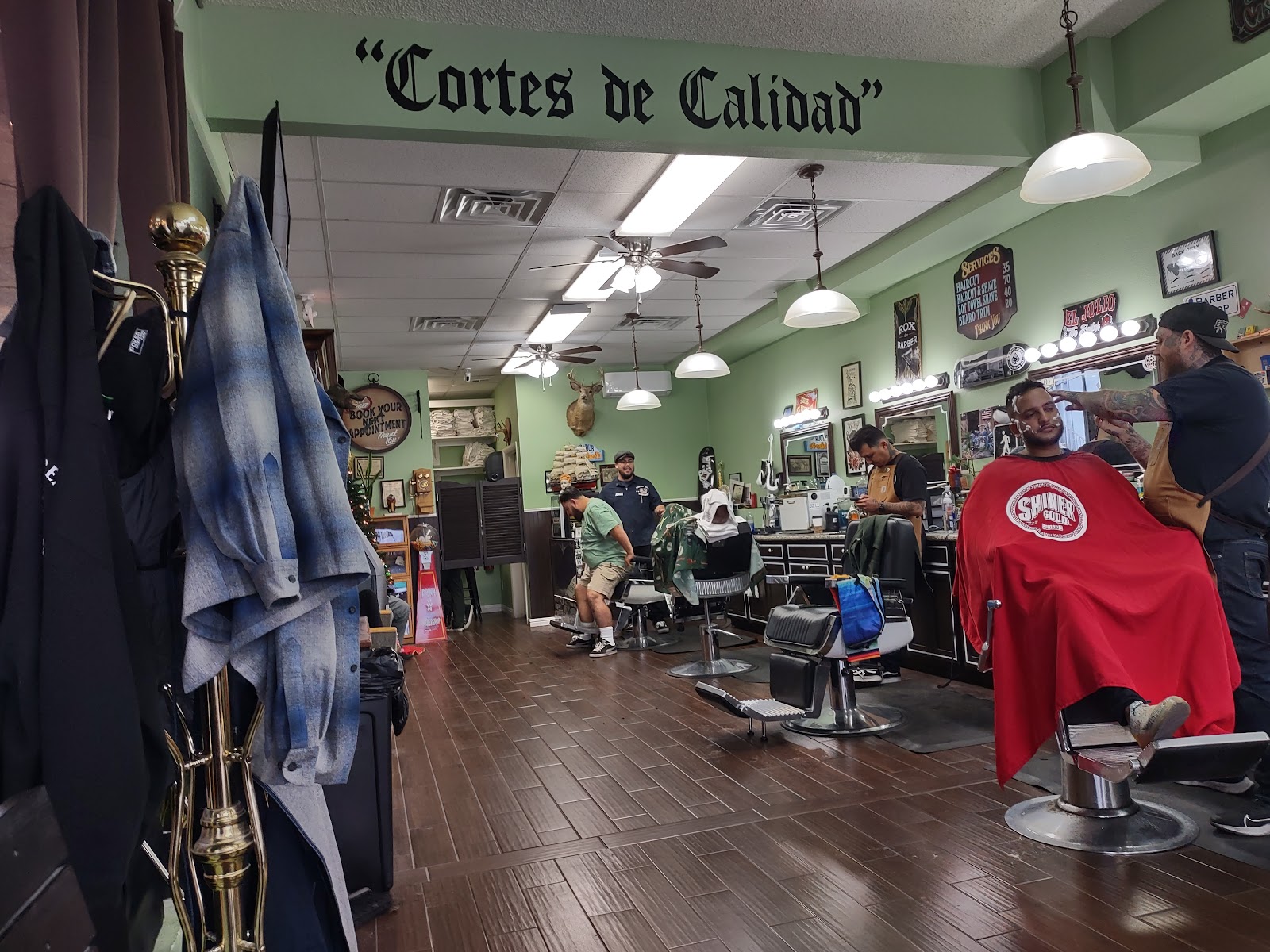 Busy barbershop interior with Cortes de Calidad wall text, several barbers and clients